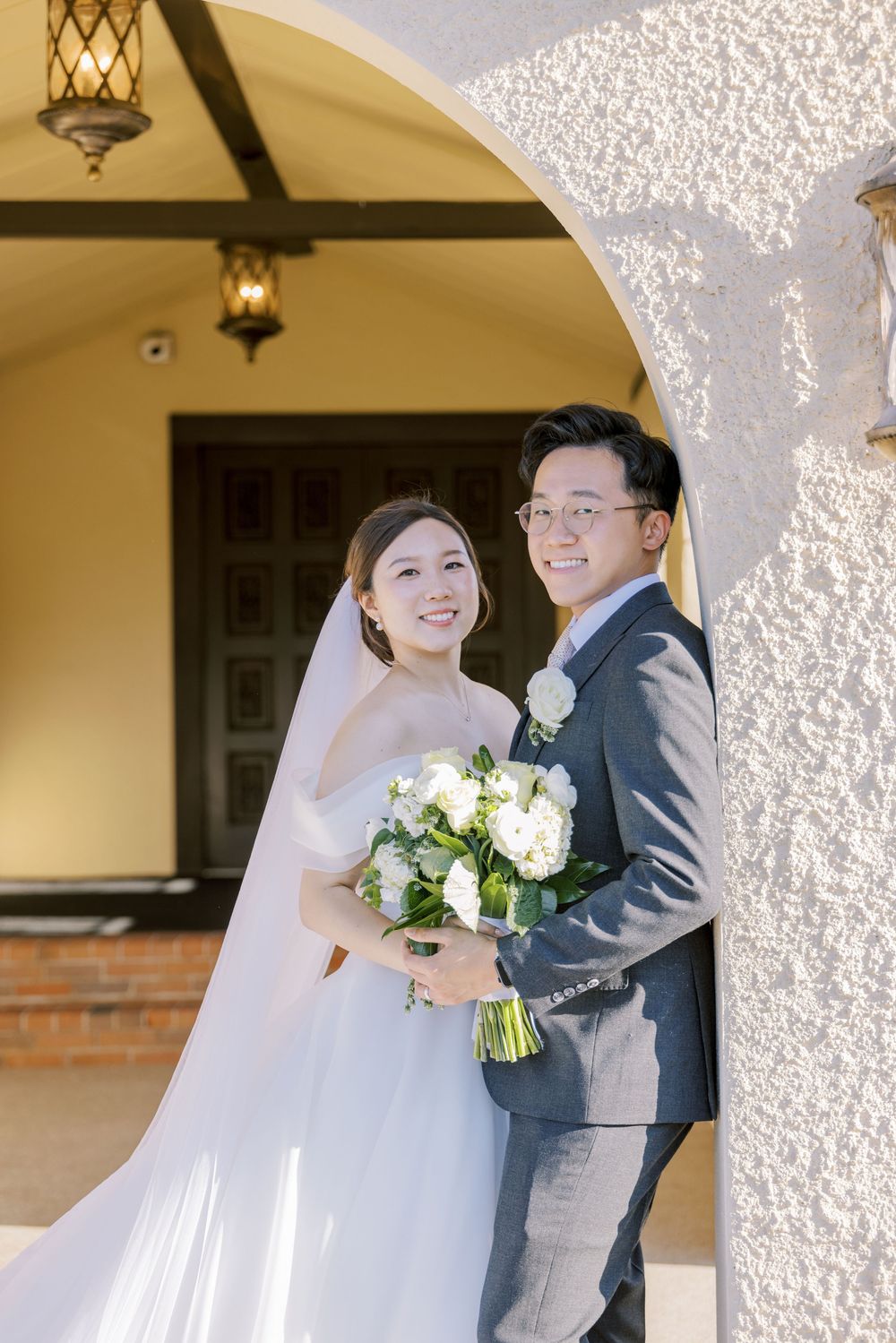 A bride and groom pose under an arched entryway with hanging lanterns while holding a white floral bouquet.