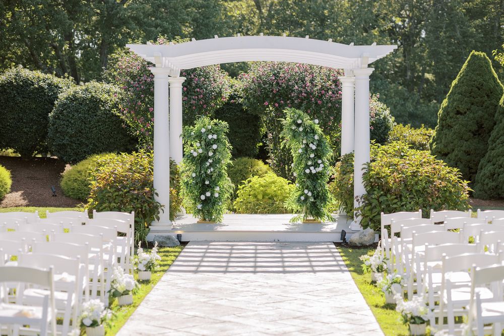 White chairs and a pergola set up for an outdoor garden wedding ceremony with lush greenery in the background.