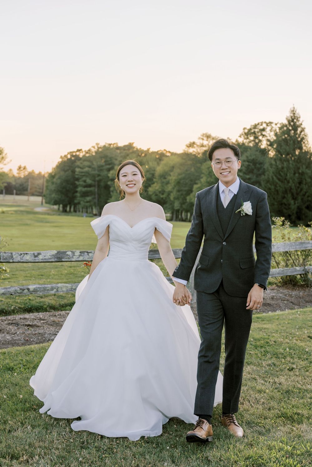 A couple walks together on grassy terrain during sunset with stone walls and trees in the background.