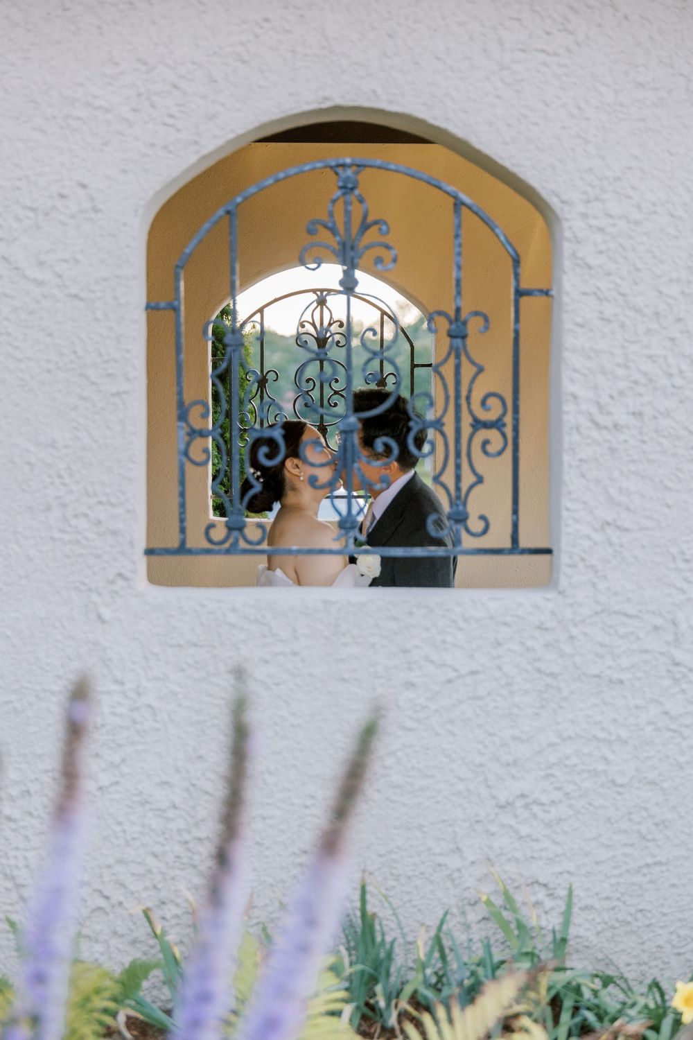A series of decorative arched windows with blue wrought iron details on a white stucco wall above flowering plants.