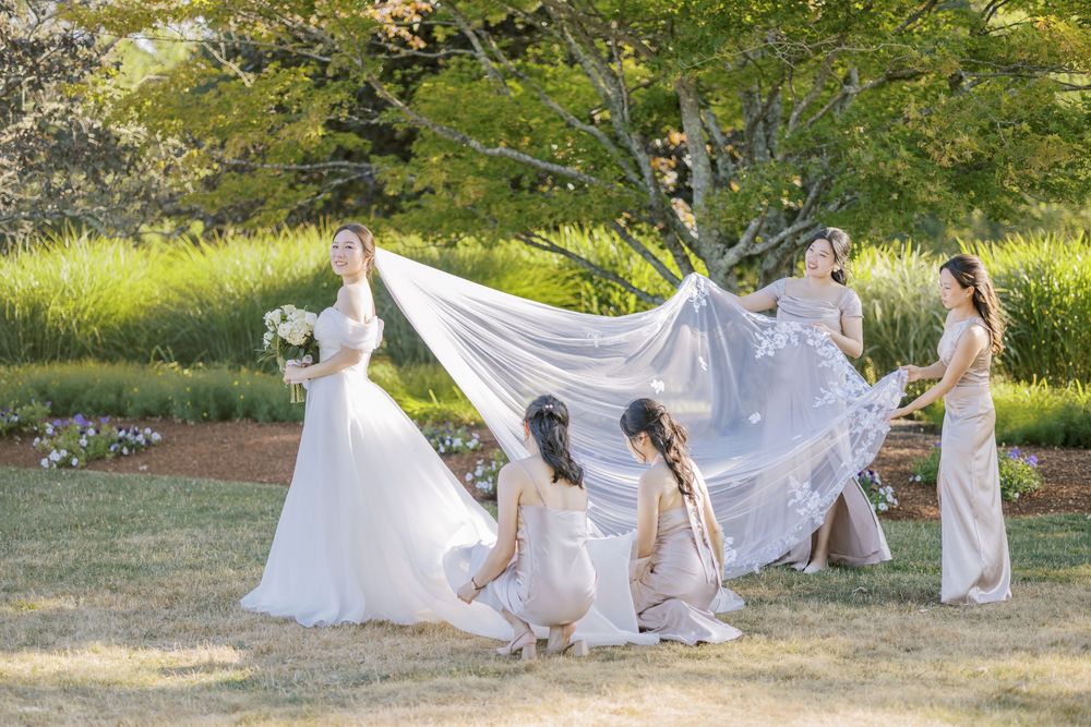 A flowing white wedding veil billows in the breeze as people gather in a sunny garden setting.