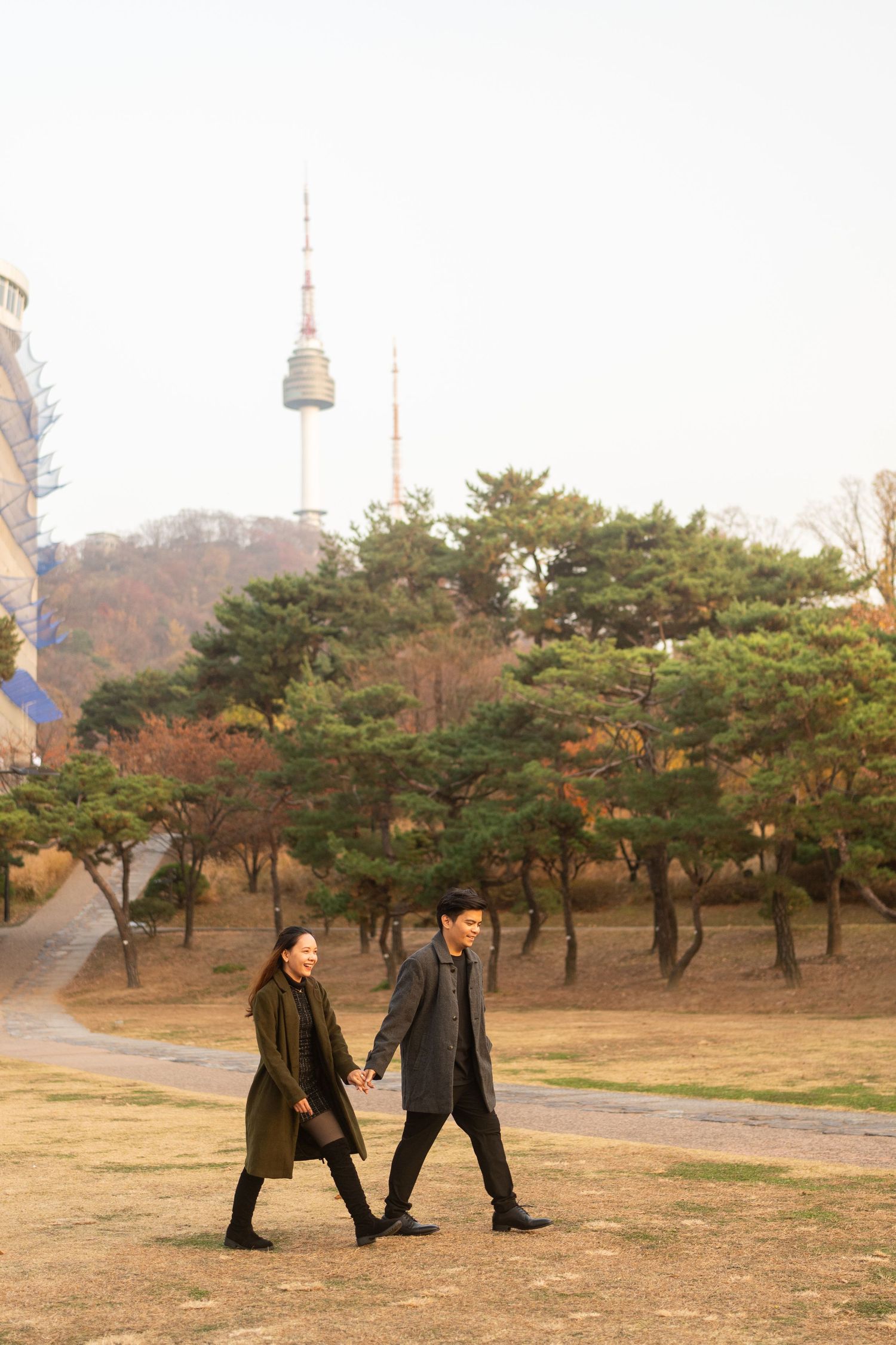 Three people walk together through a park with pine trees and a tower visible in the background on an autumn day.