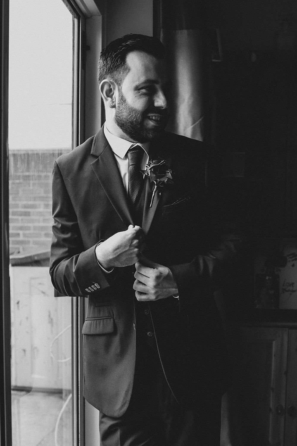 A black and white photo of groom in his suit adjusting his tie while standing near windows.