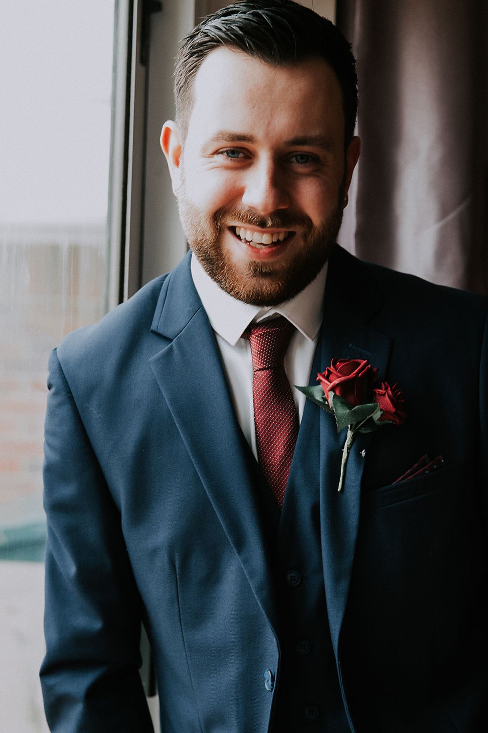 A groom in a navy suit and burgundy tie smiles while standing near a window.