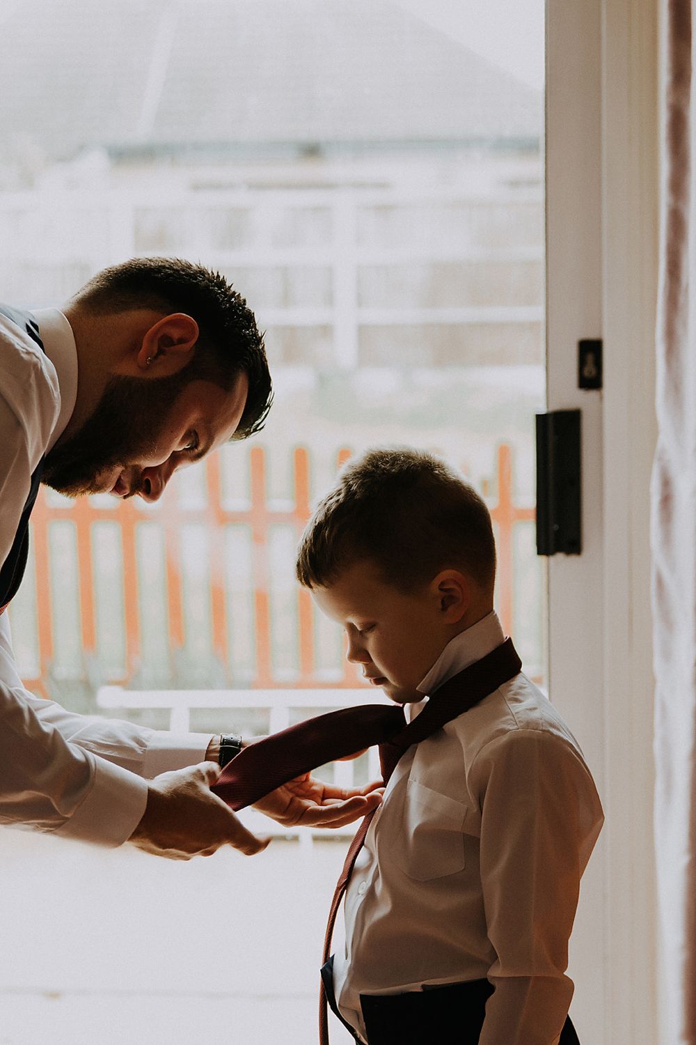 groom and his son standing by a bright window fixing his sons tie