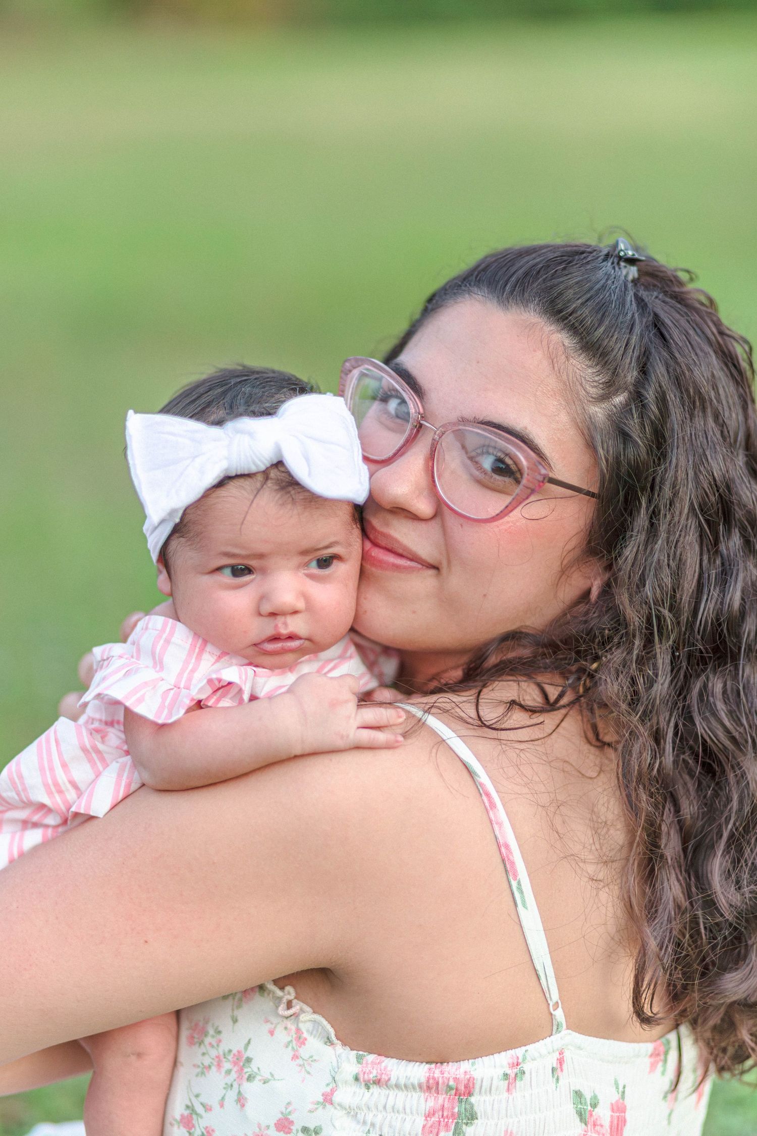 Mother with pink glasses kissing her newborn baby girl wearing a white bow headband outdoors.