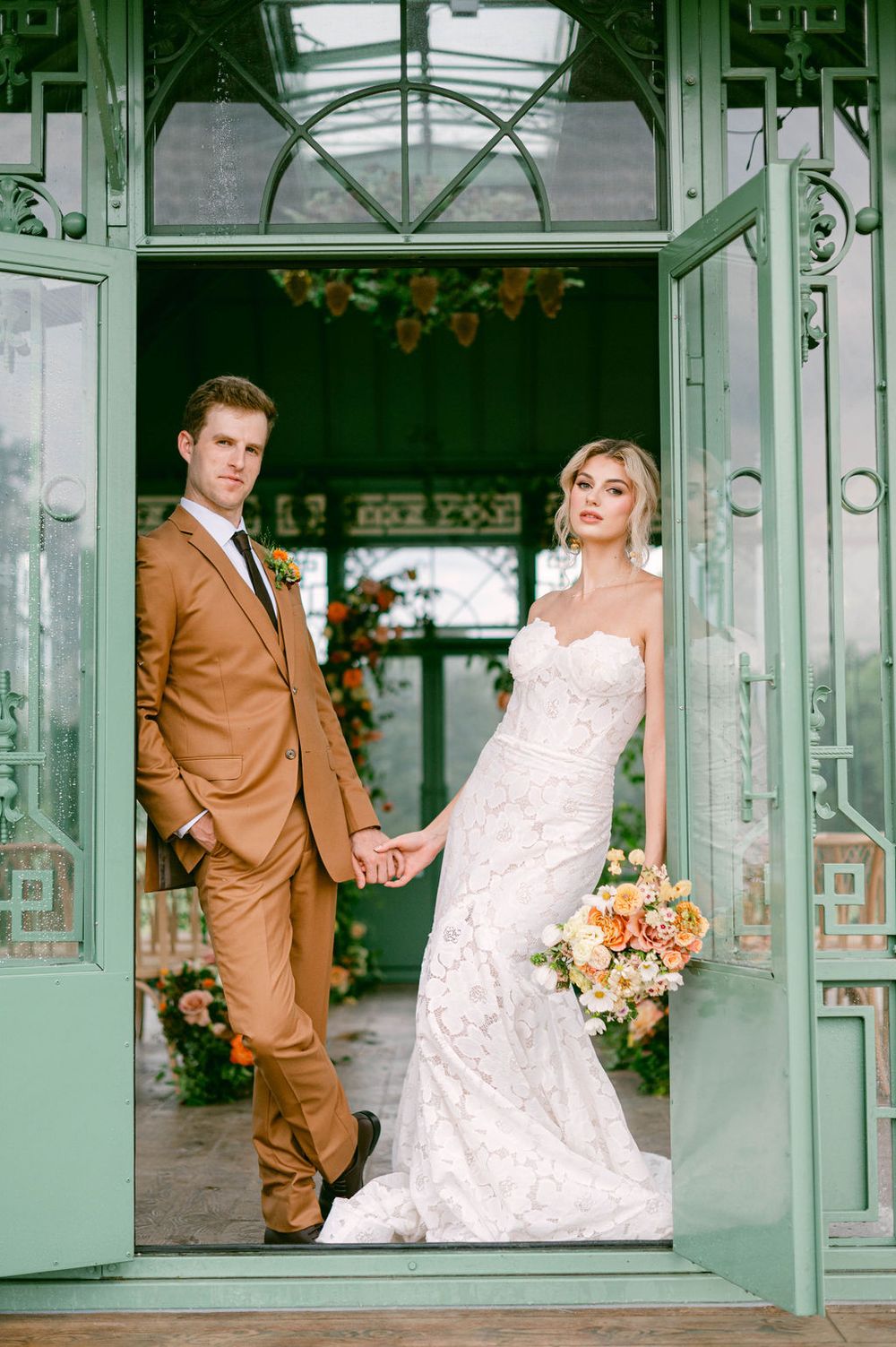 A bride in a lace gown and groom in a brown suit pose in a vintage mint green doorway with floral accents.