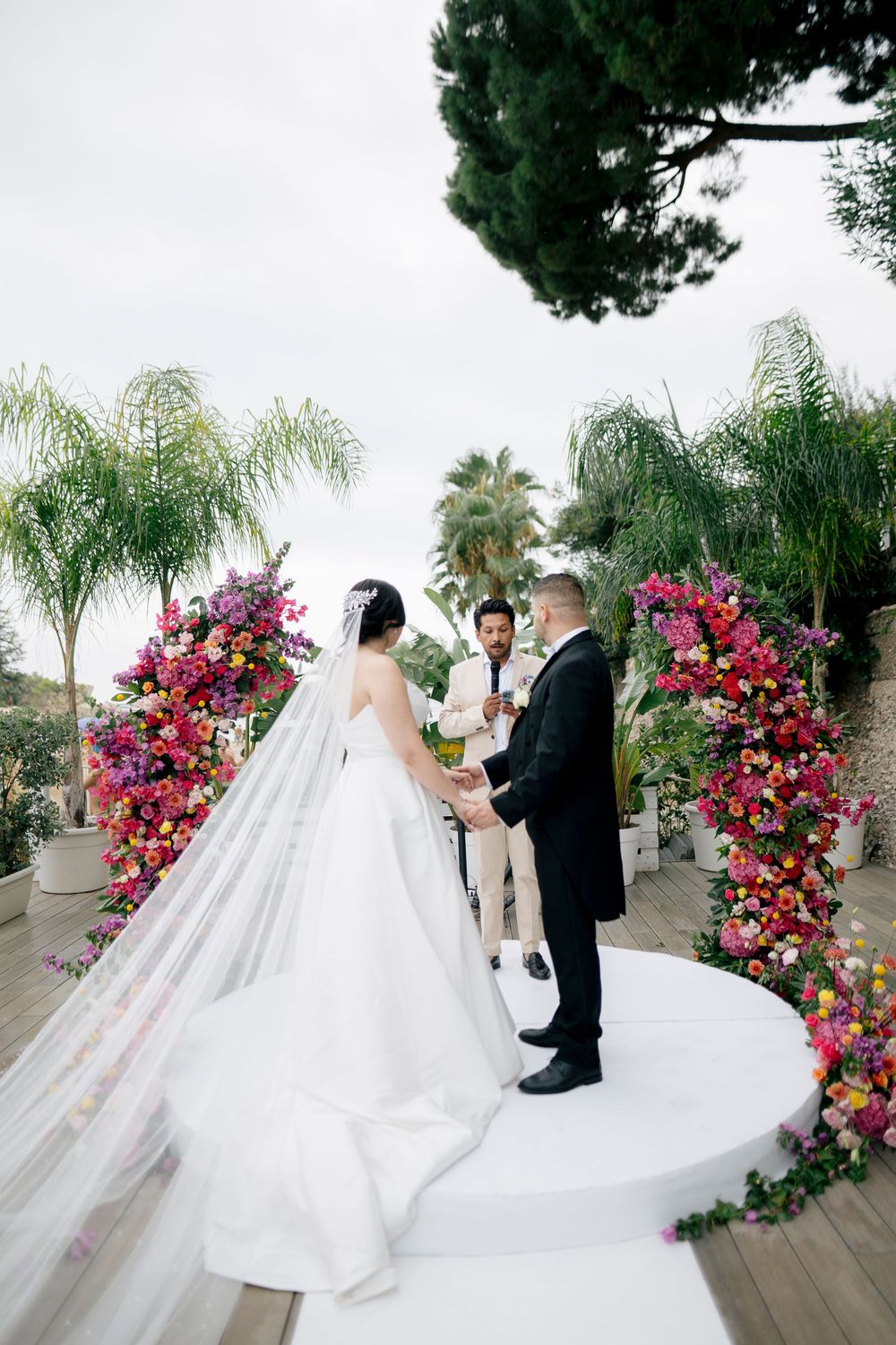 A romantic wedding ceremony scene with vibrant pink floral arch and flowing white gown against a palm tree backdrop.