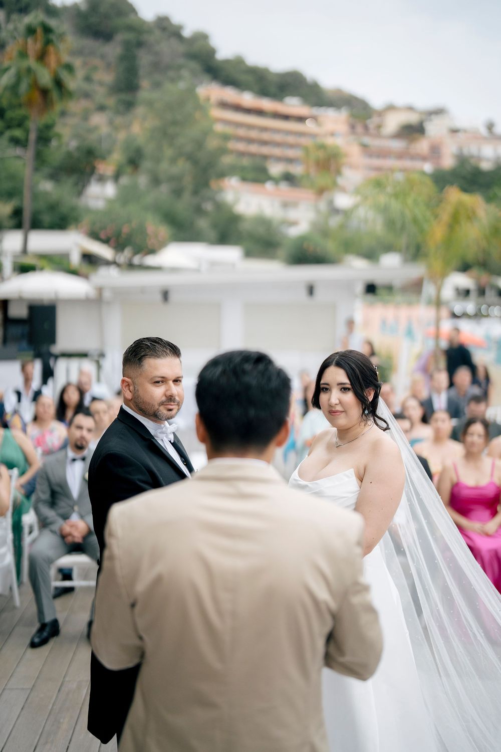 Wedding guests line up along a wooden deck overlooking water and buildings during an outdoor celebration.