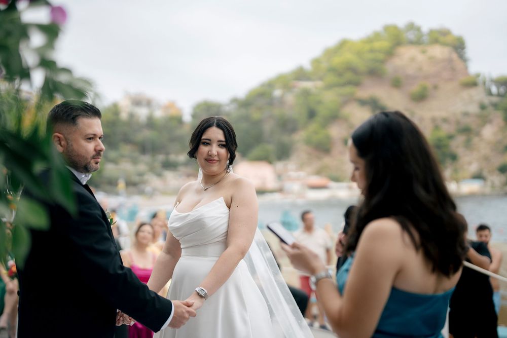 Wedding ceremony guests gather outdoors at a scenic beach location with mountains in the background.