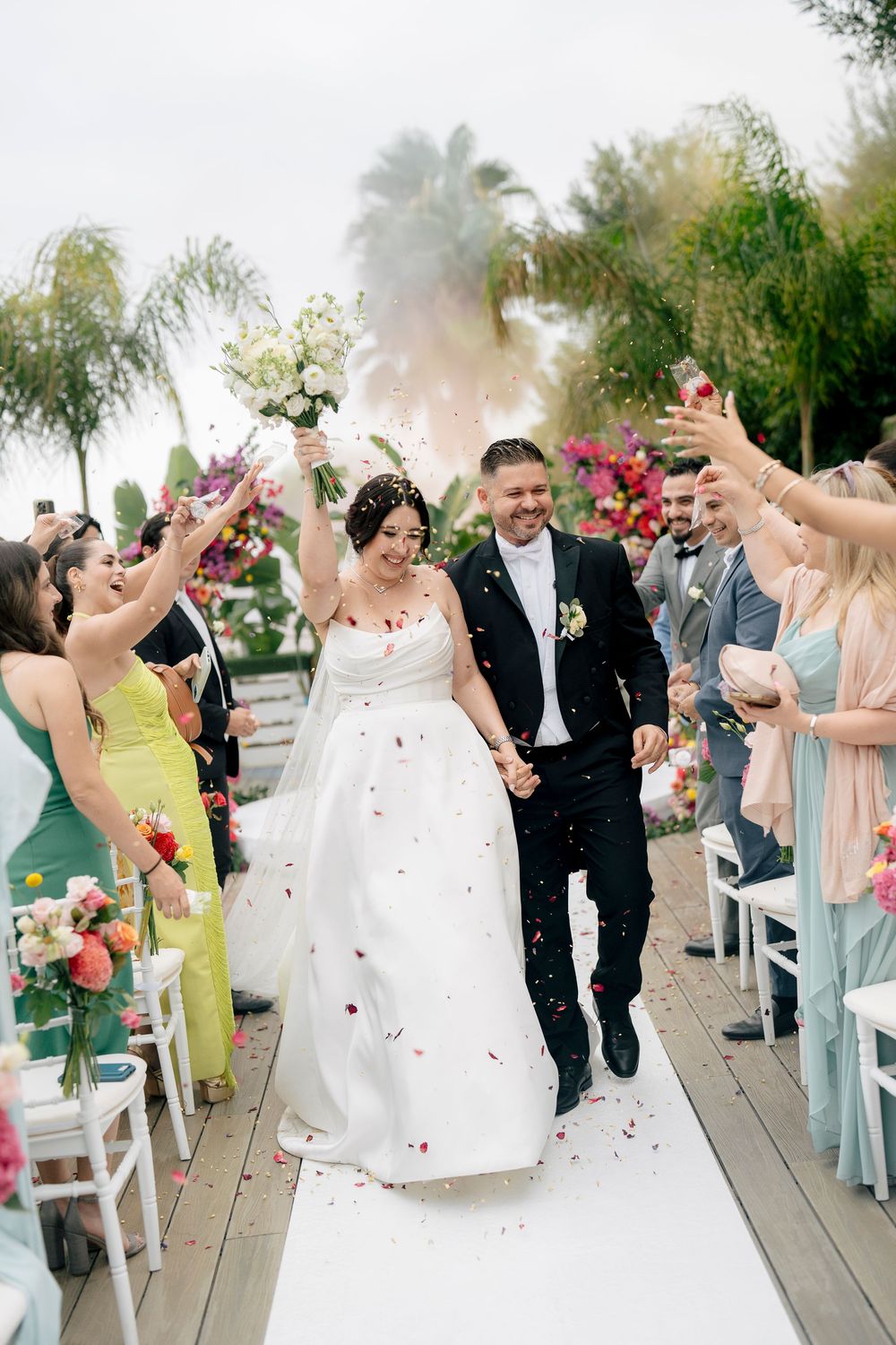Wedding guests toss flower petals as newlyweds walk down the aisle at an outdoor ceremony.