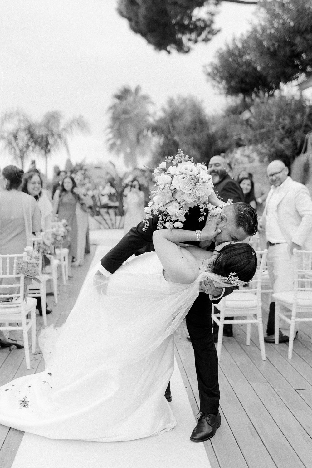 A romantic dip kiss between newlyweds on an outdoor deck with white chairs and guests in the background.