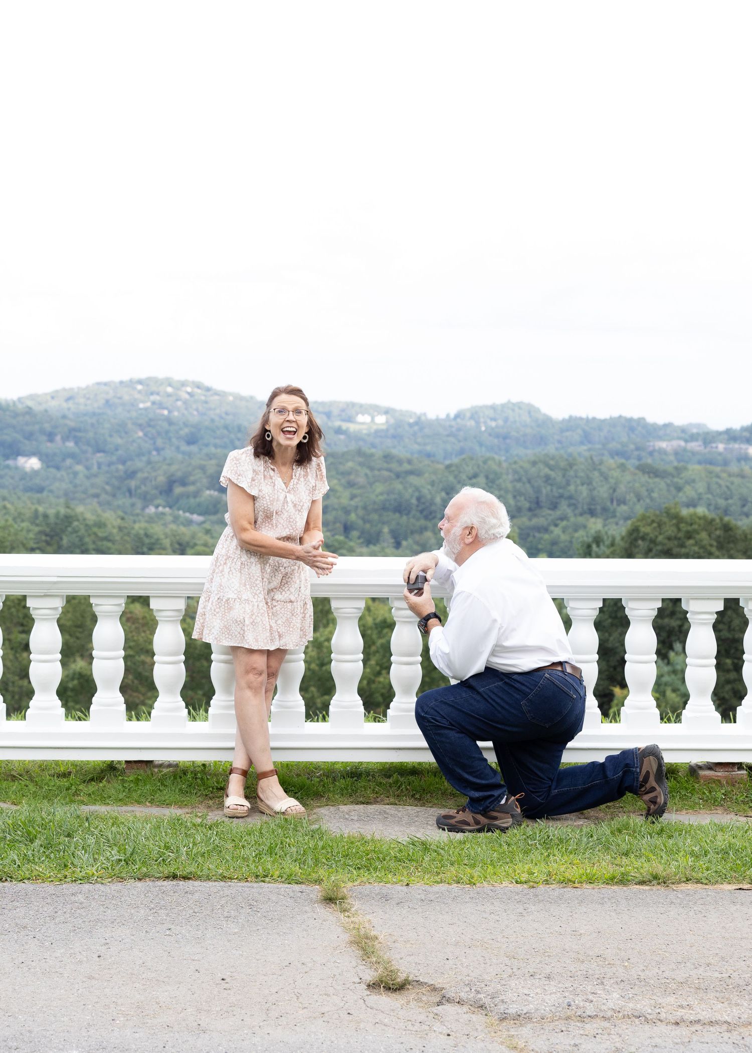 Man kneeling on one knee proposing to a surprised woman on a terrace with mountain views.