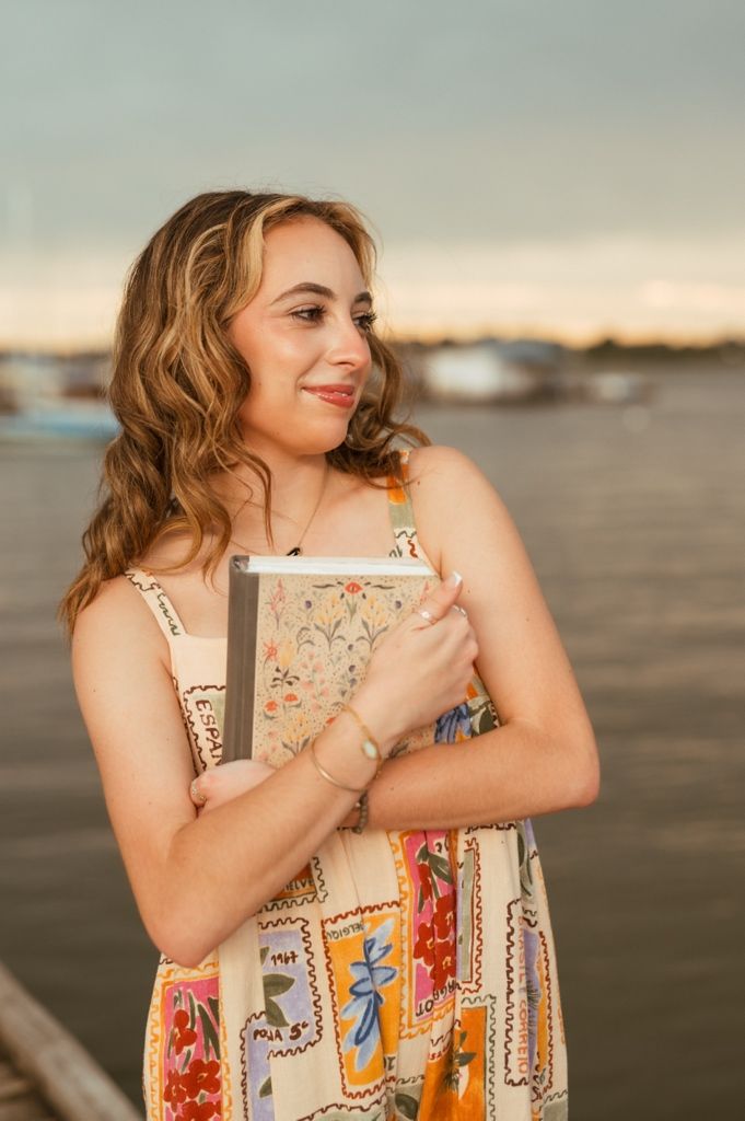 Woman wearing vintage-style printed dress hugs herself while posing on beach at golden hour.