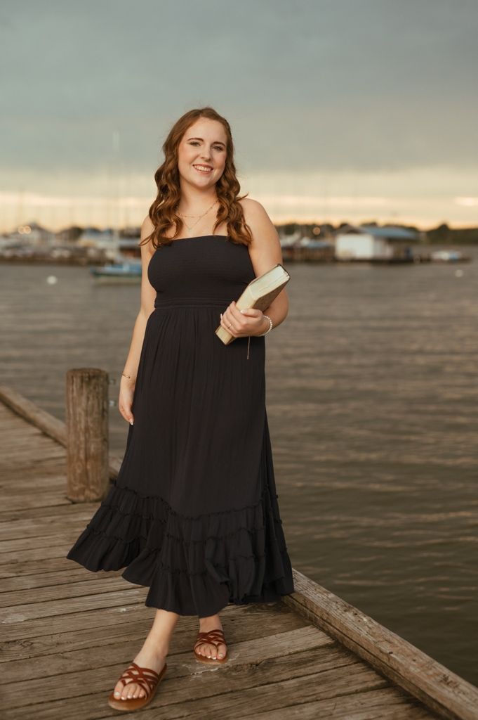 Woman in flowing black dress sits casually on wooden pier overlooking waterfront at dusk.