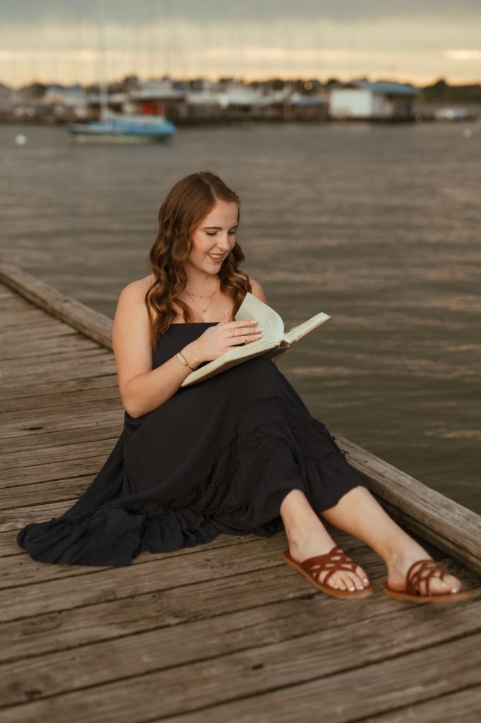 Person in black dress reading while relaxing on wooden dock at sunset.