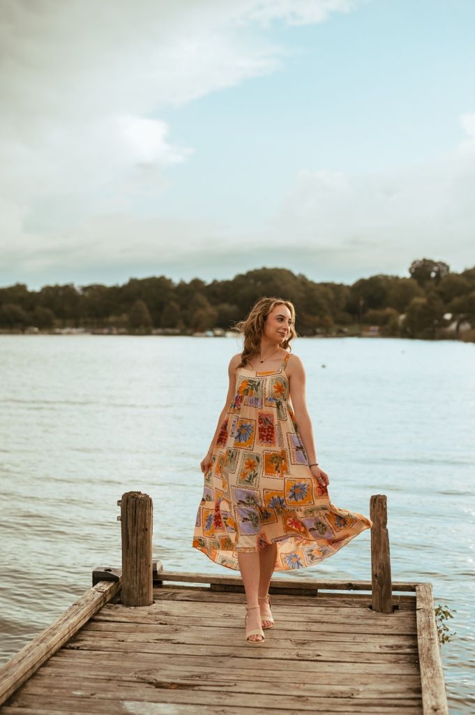 Woman in floral sundress twirling on wooden dock by lake during sunset.
