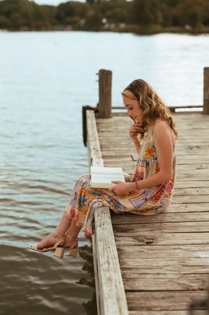 Person reading book while sitting on edge of wooden dock over peaceful lake.