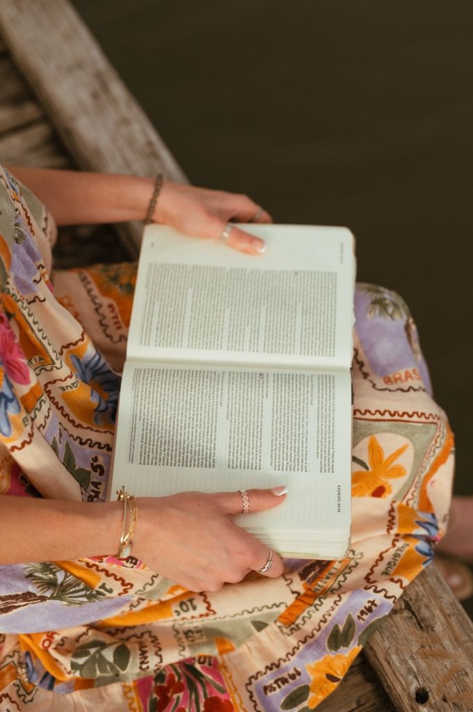 Close-up of hands holding open book on wooden dock with floral dress visible.