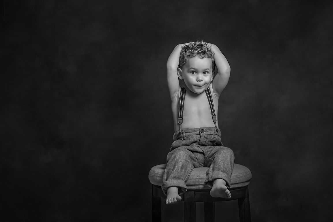 Black and white portrait of a toddler boy sitting on a stool with suspenders.