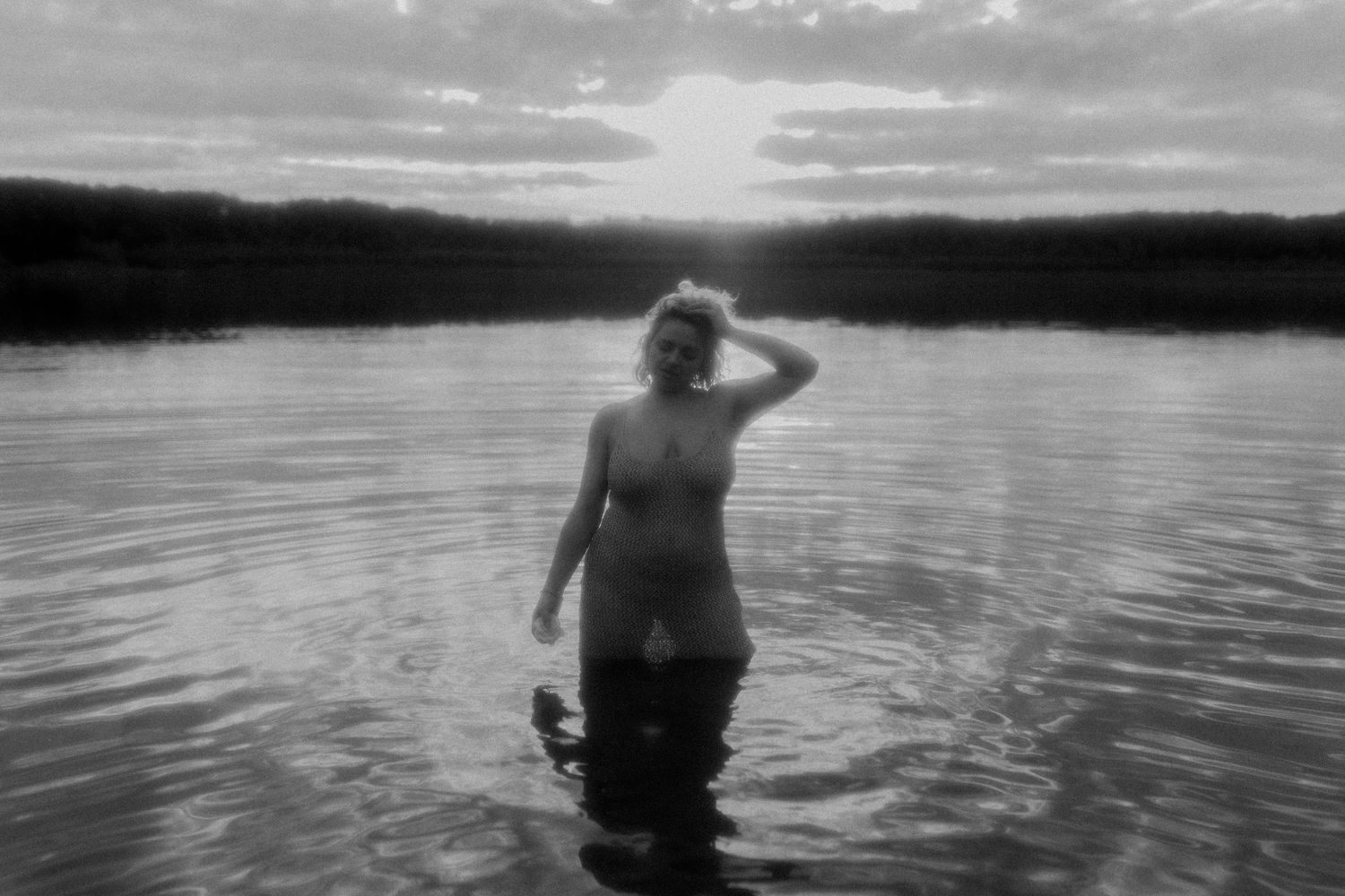 A black and white panoramic sequence shows a figure wading deeper into a tranquil lake at sunset.