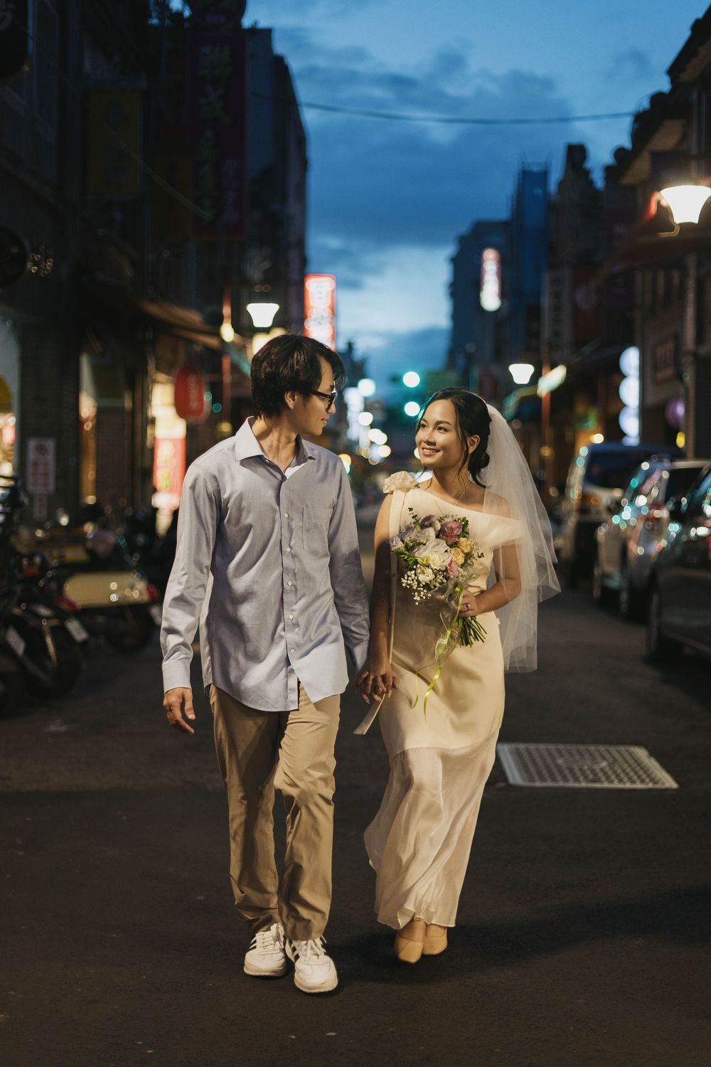 Couple in wedding attire walking hand-in-hand on a city street at night, bride holding bouquet.