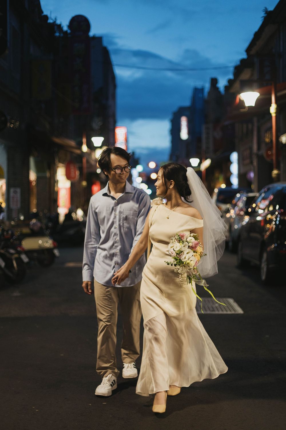 Newlyweds strolling along a city street at dusk, bride in white gown holding a floral bouquet.