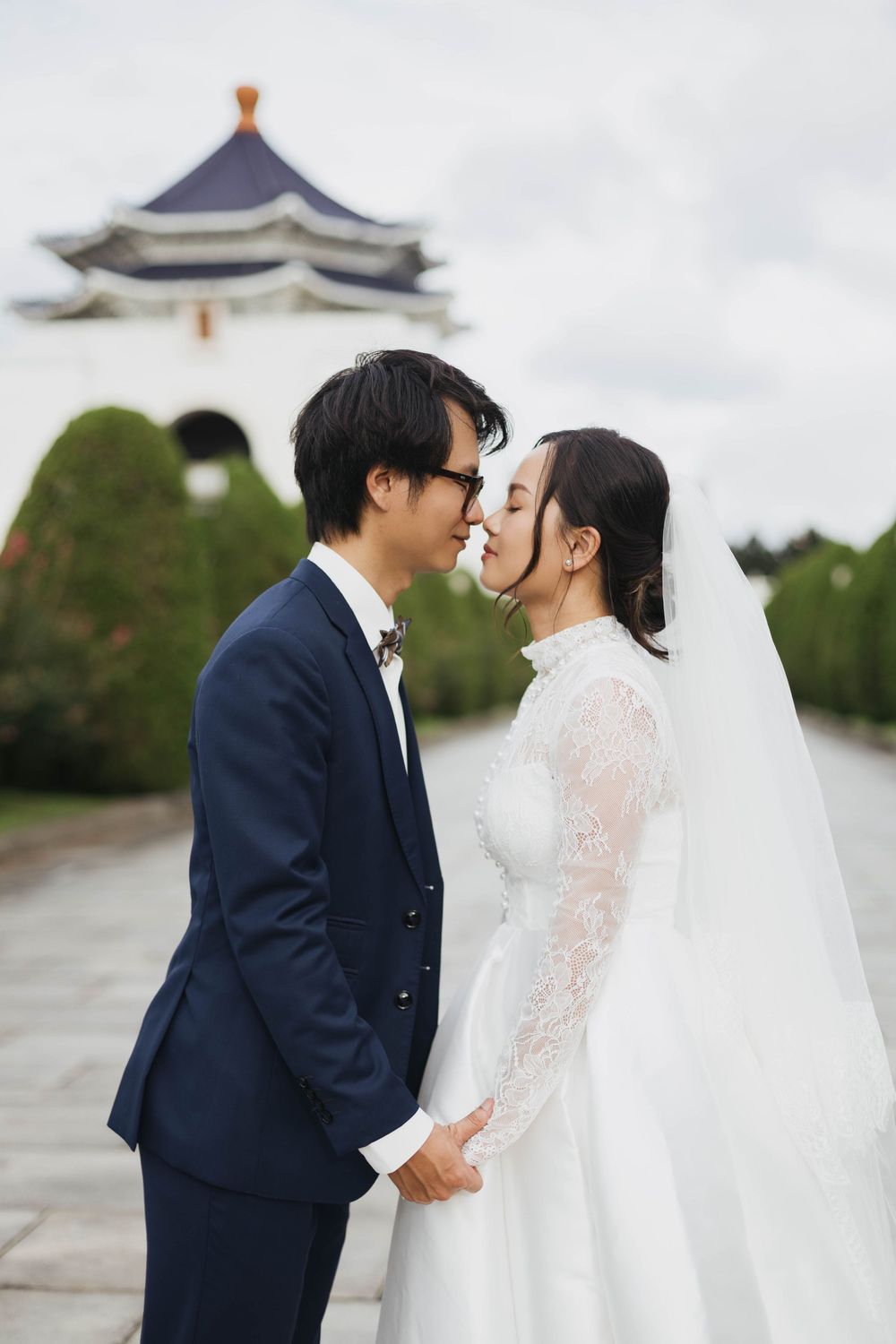 Couple sharing a romantic nose-to-nose moment in wedding attire near a traditional Asian monument.