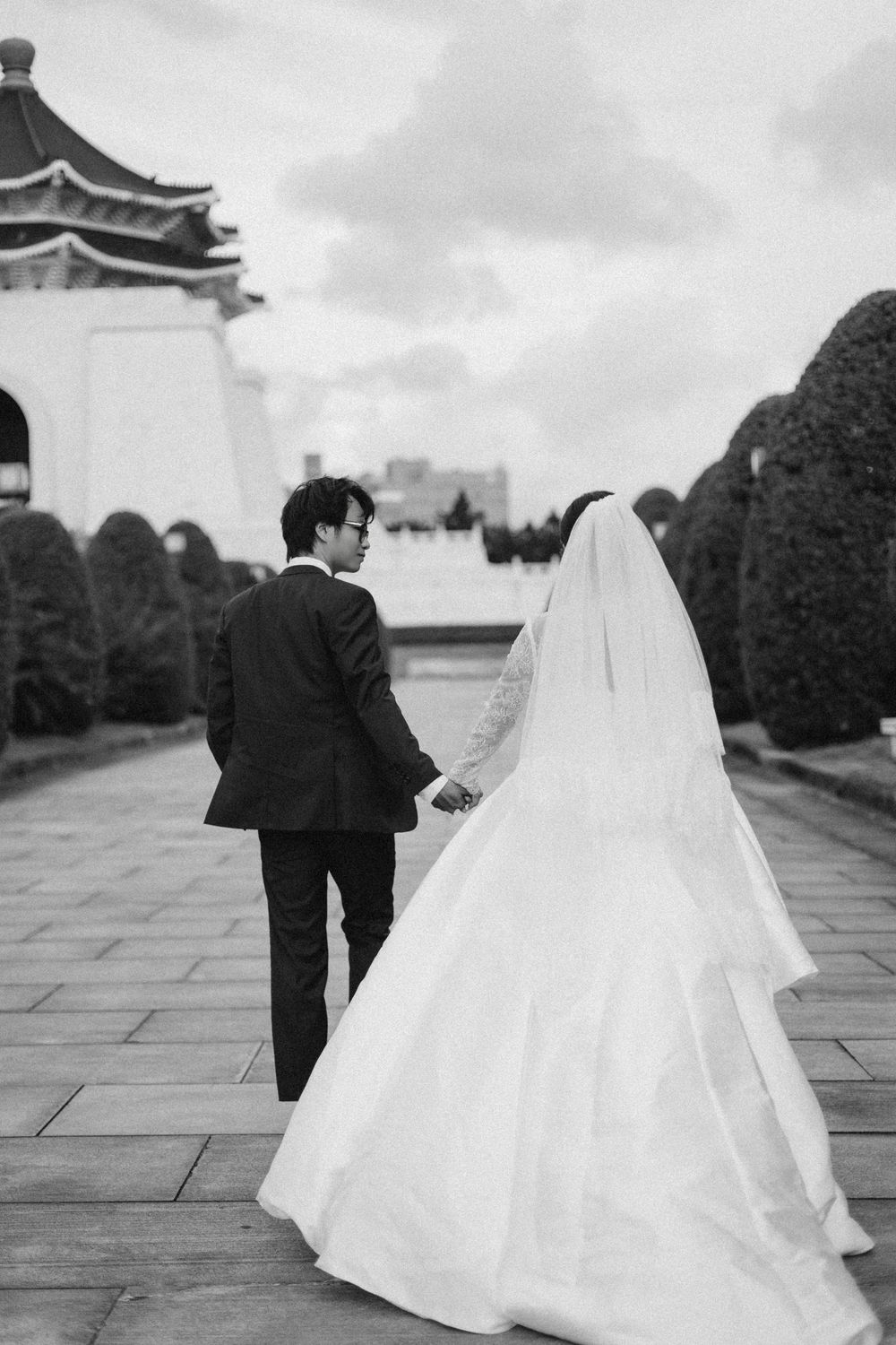 Black and white photo of newlyweds walking hand-in-hand toward a traditional Asian pavilion.