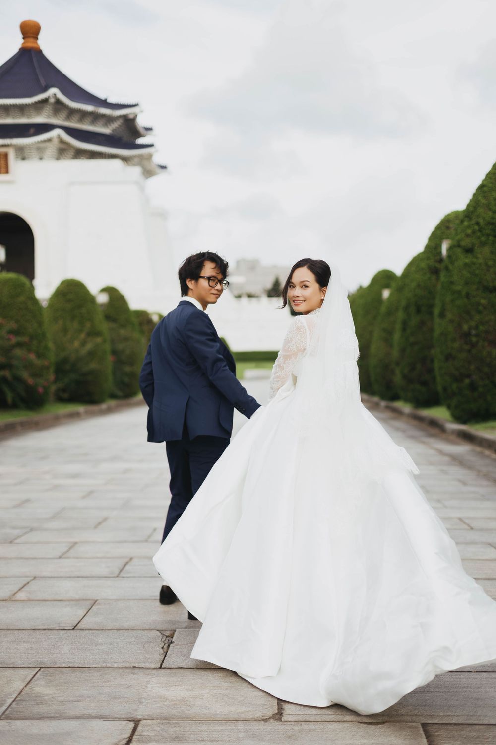 Bride in flowing white gown glancing back while holding groom's hand near an Asian monument.