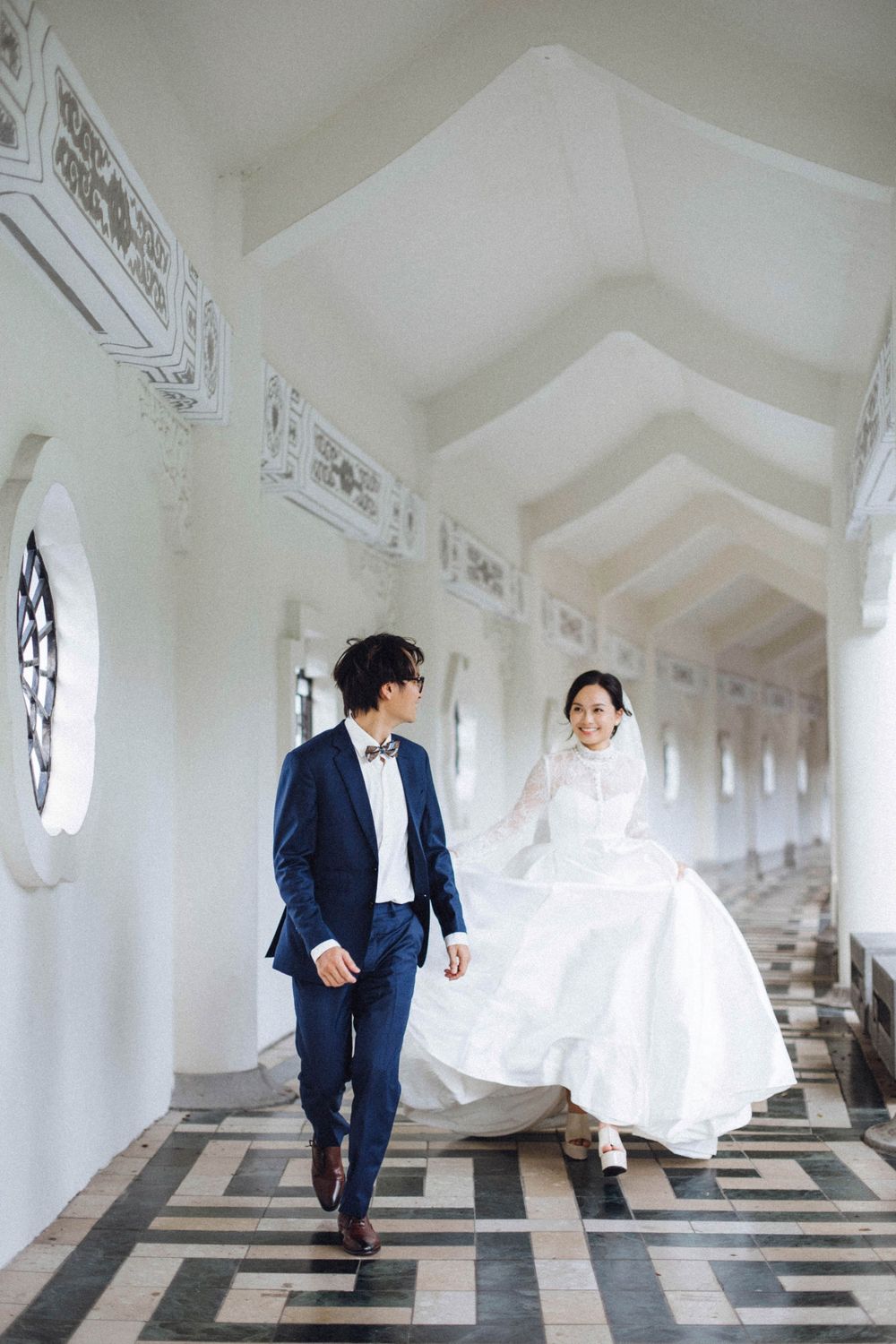 Happy couple strolling through a grand white arched hallway, bride lifting her flowing white dress.