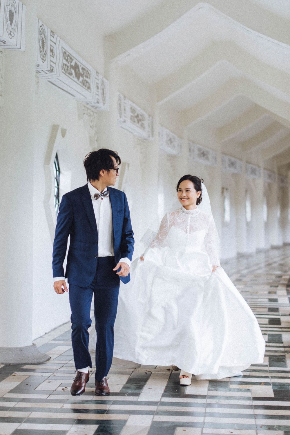 Bride in lace wedding gown and groom in navy suit walking hand in hand through an elegant white corridor.