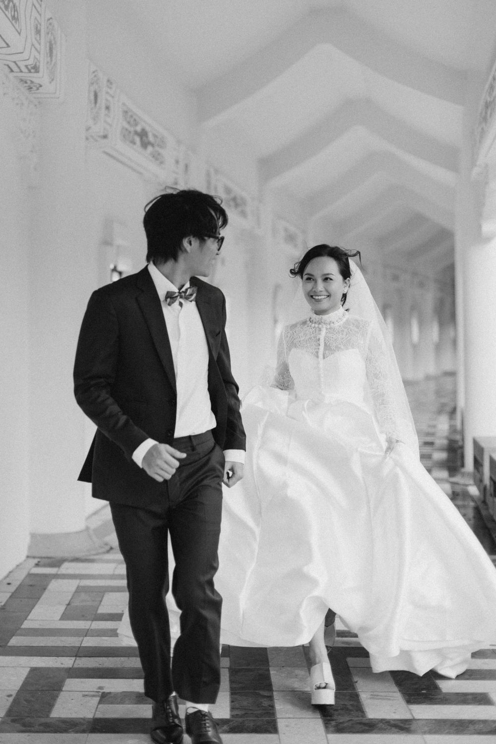 Black and white photo of smiling bride and groom walking through an ornate columned corridor.