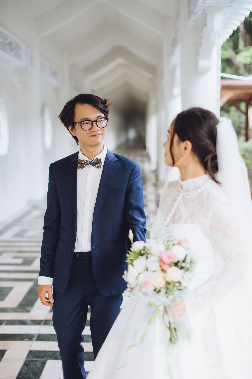 Groom in navy suit with bow tie smiling at bride in lace wedding dress in elegant corridor.