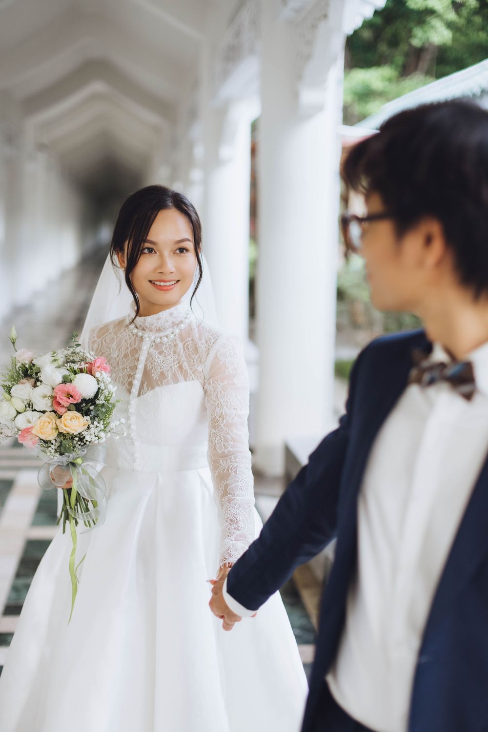 Smiling bride in lace wedding gown holding bouquet while holding groom's hand in white corridor.