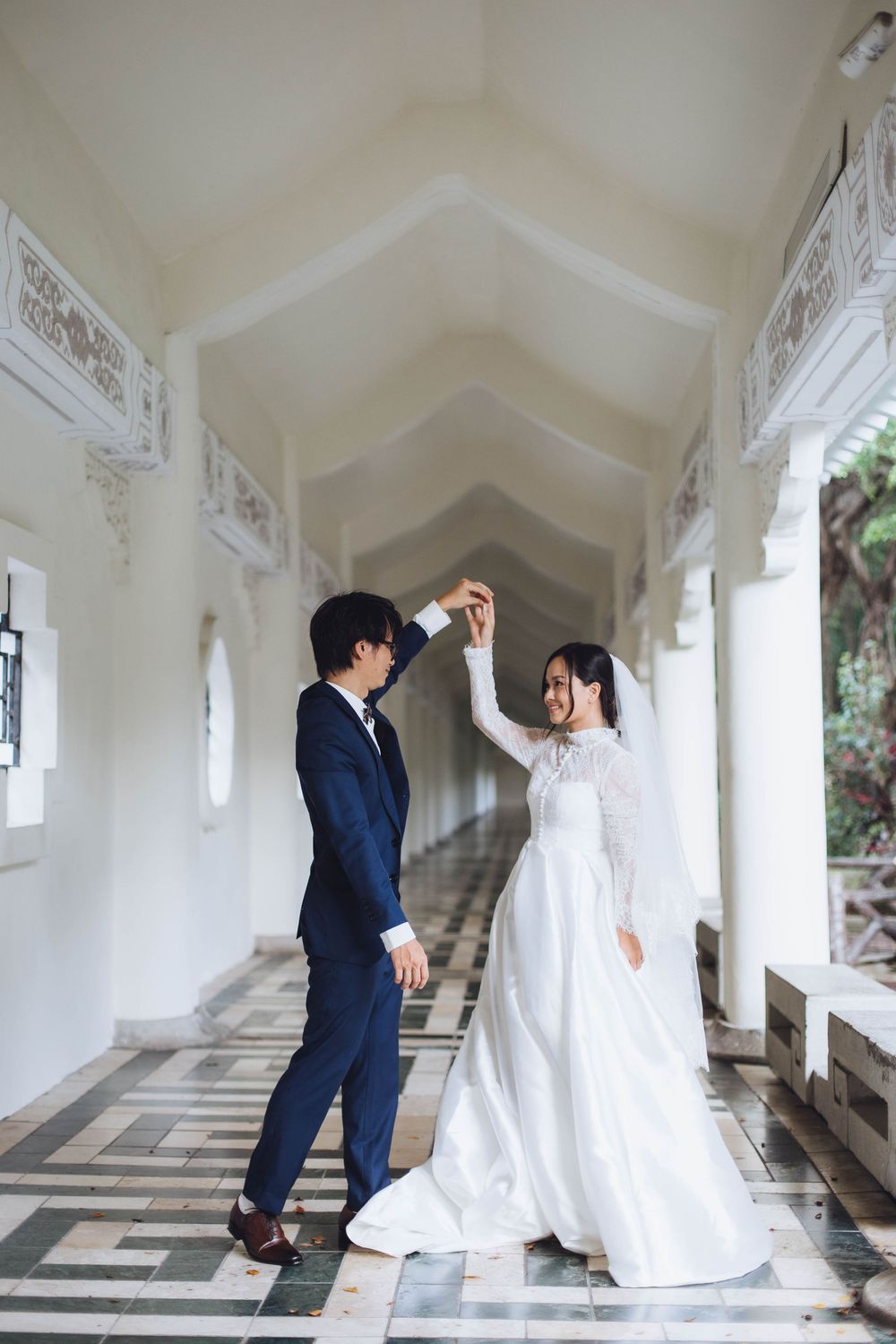 Couple dancing joyfully in a bright white arched corridor, bride in lace gown with veil.
