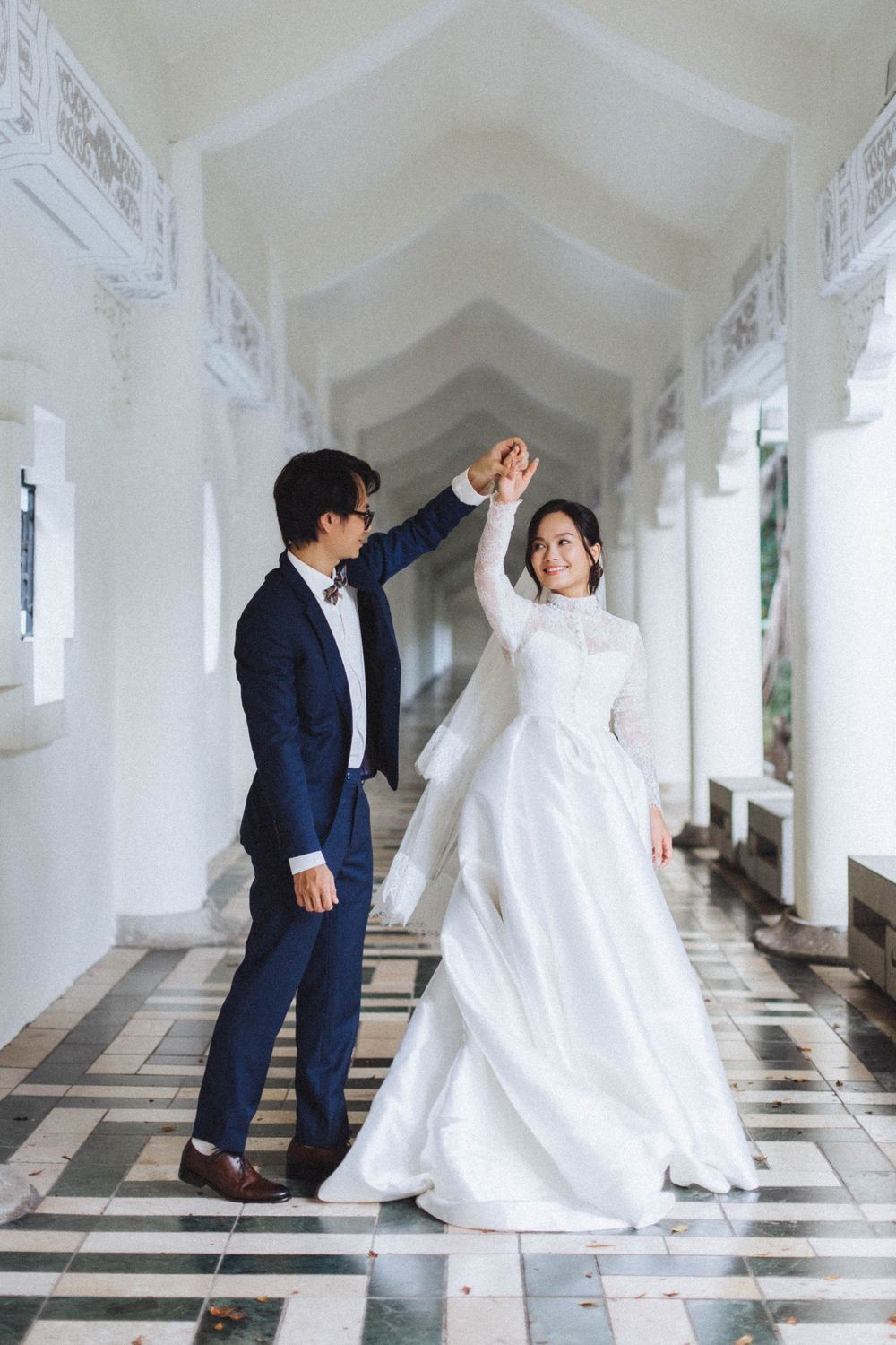 Groom in navy suit spinning bride in elegant white lace gown in a beautiful white arched hallway.