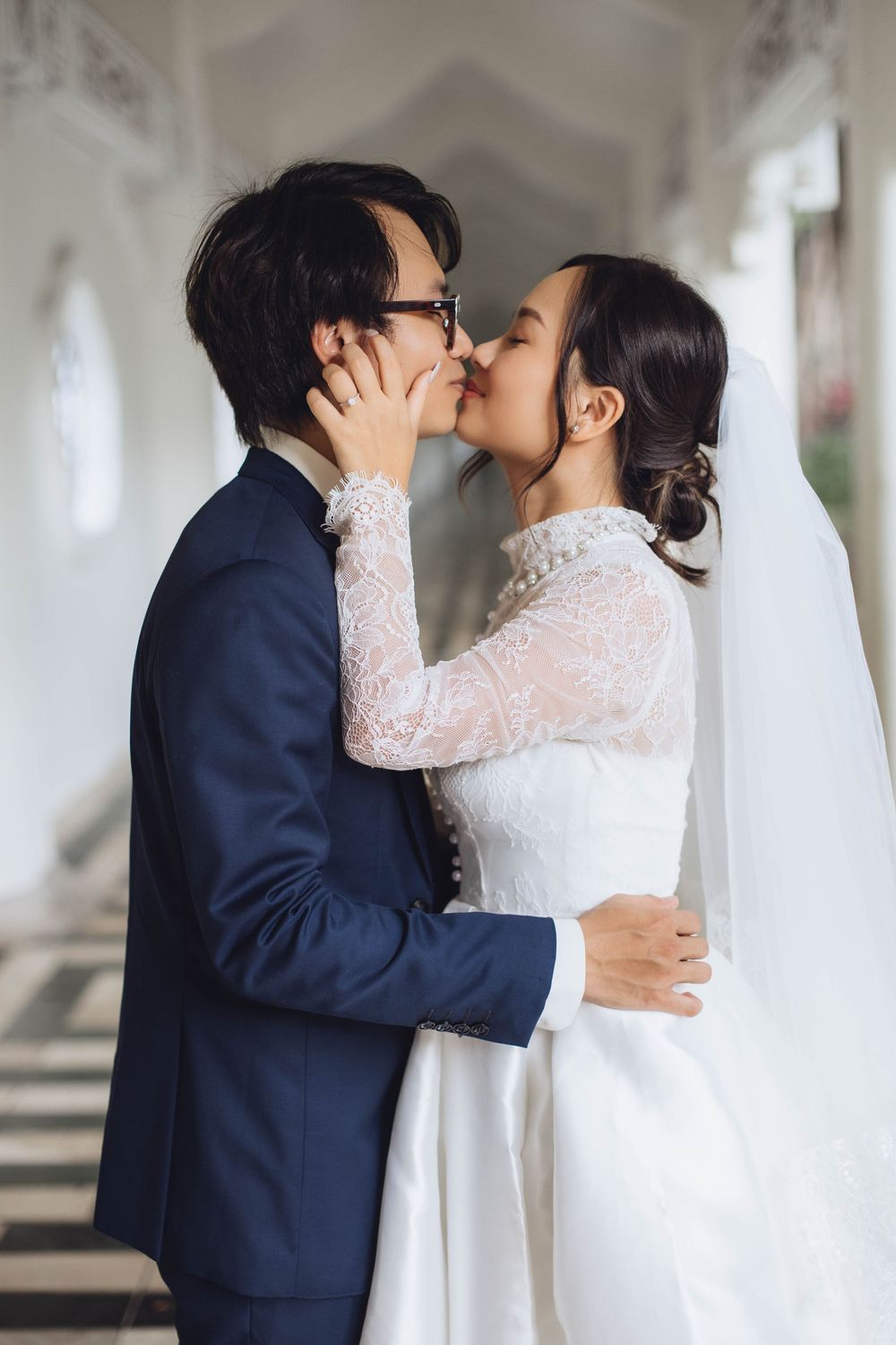 Couple sharing a passionate kiss in a white arched corridor, bride in lace wedding dress.