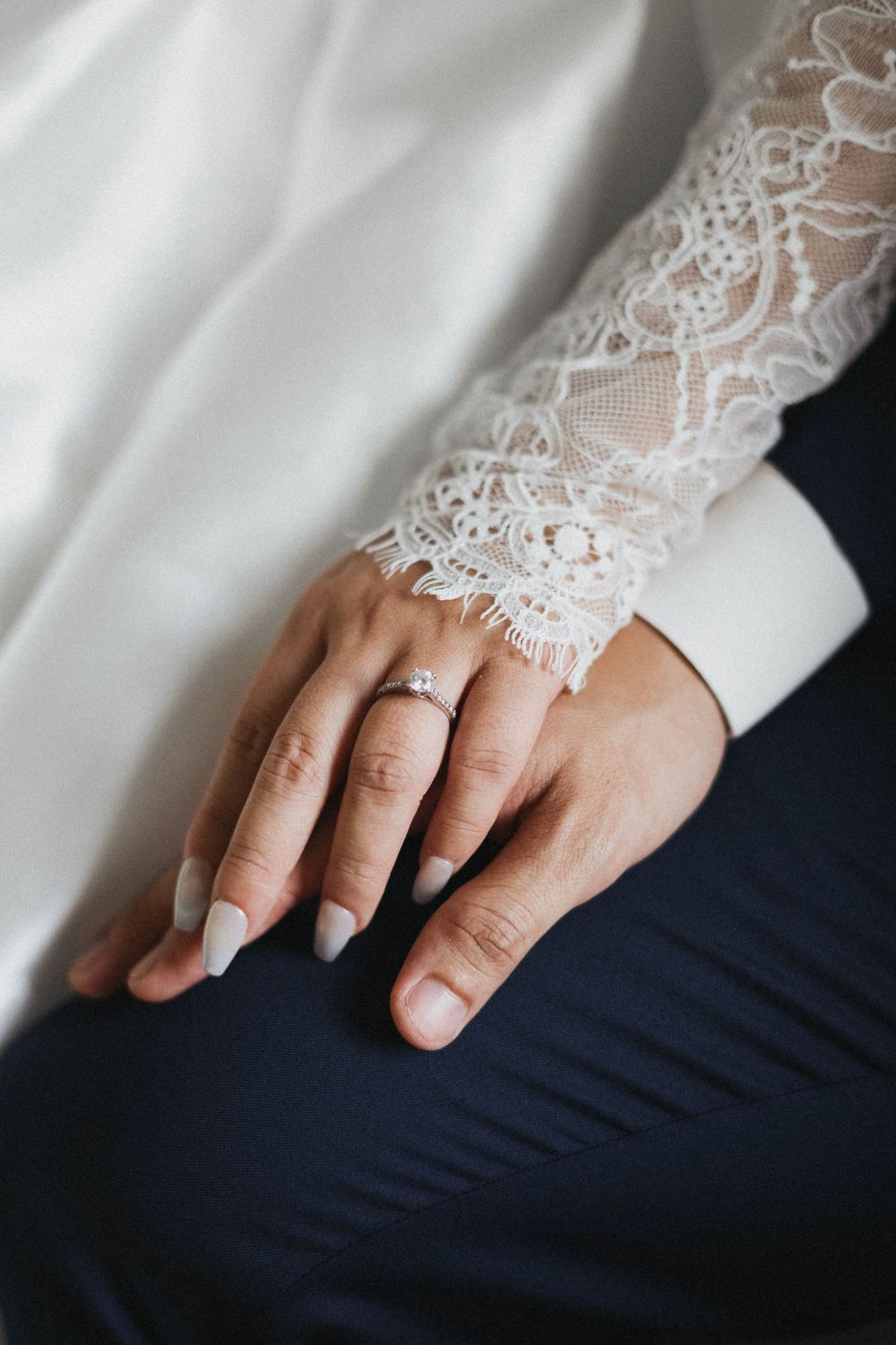 Close-up of bride's hand with diamond engagement ring resting on groom's navy suit trousers.
