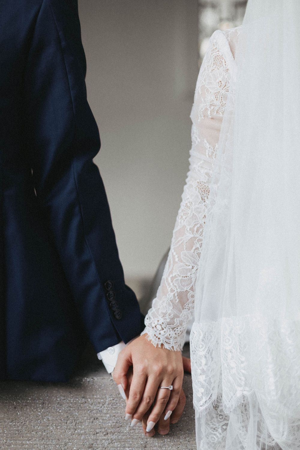 Close-up of bride and groom holding hands, showcasing the bride's diamond ring and lace sleeve.