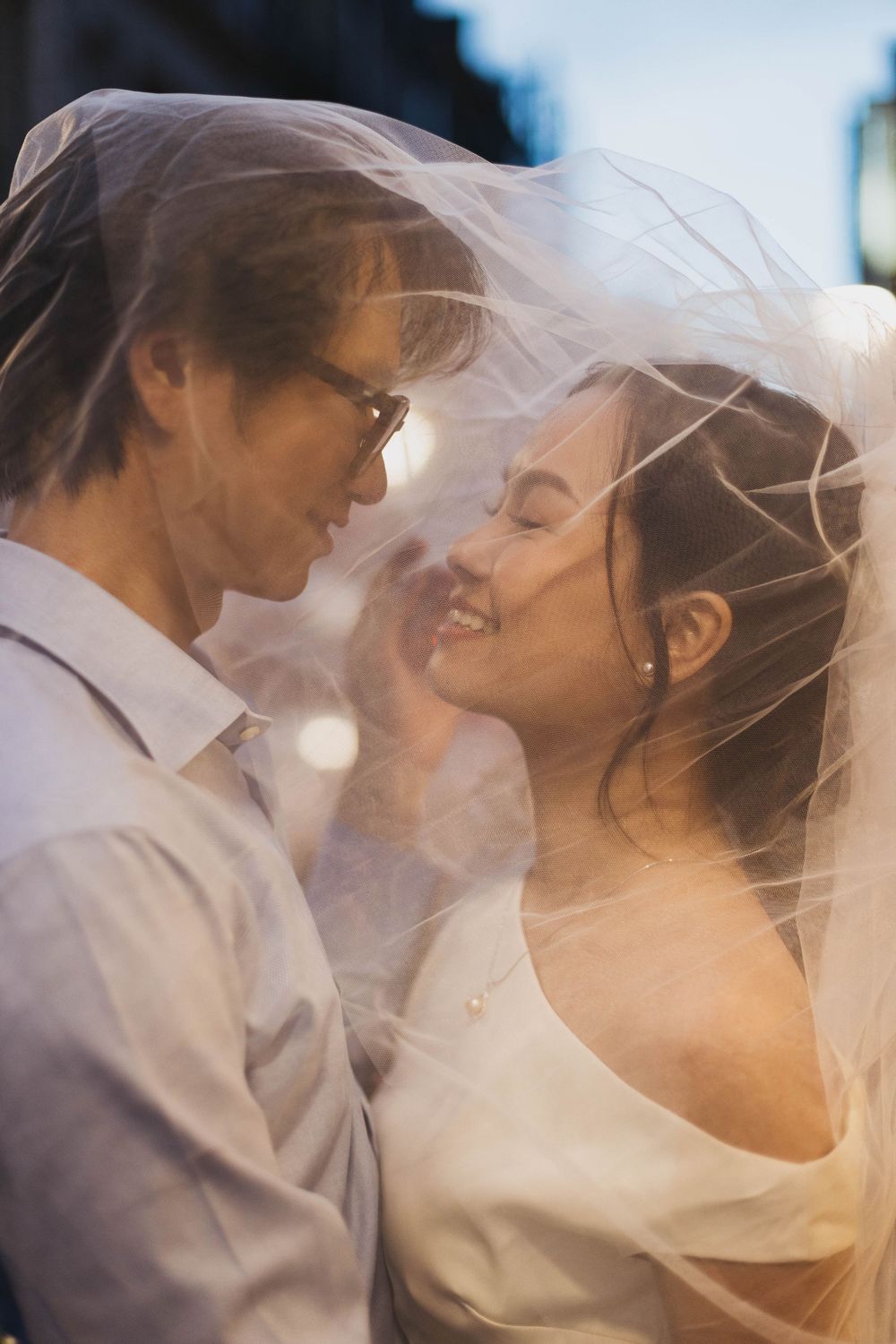 Bride and groom sharing a romantic moment under a sheer wedding veil, smiling and gazing at each other.