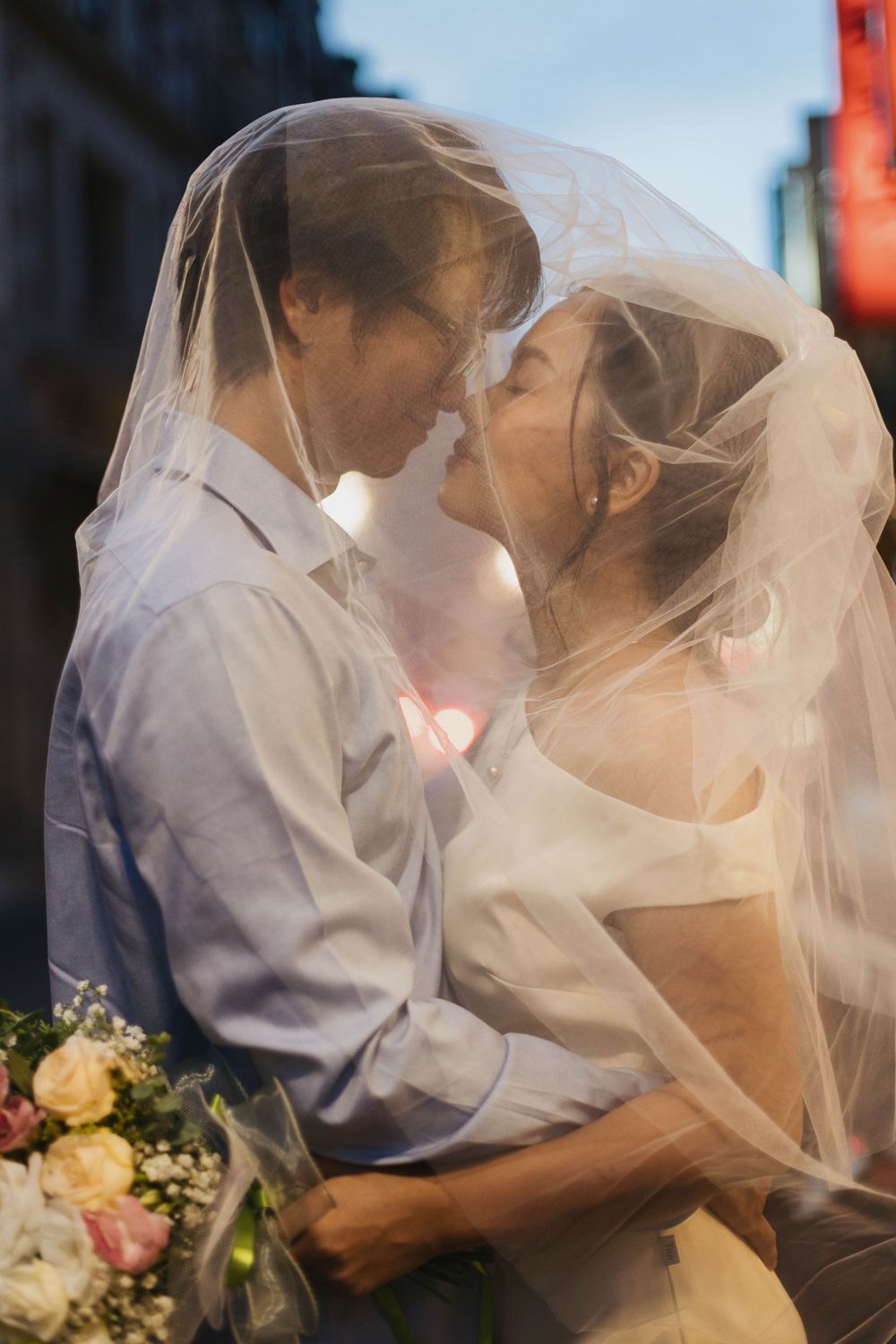 Newlyweds nearly kissing beneath a flowing bridal veil, bride holding a bouquet of pastel flowers.