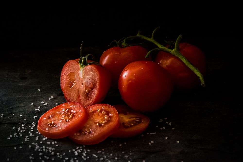 Fresh red tomatoes, some sliced in half, with scattered salt crystals on a dark surface.