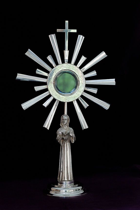 Silver Catholic monstrance with sunburst rays, cross, and praying figure on a black background.