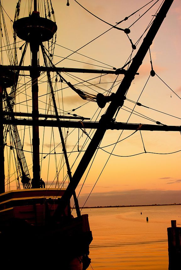 Silhouette of a tall ship's mast and rigging against a stunning orange and gold sunset sky.
