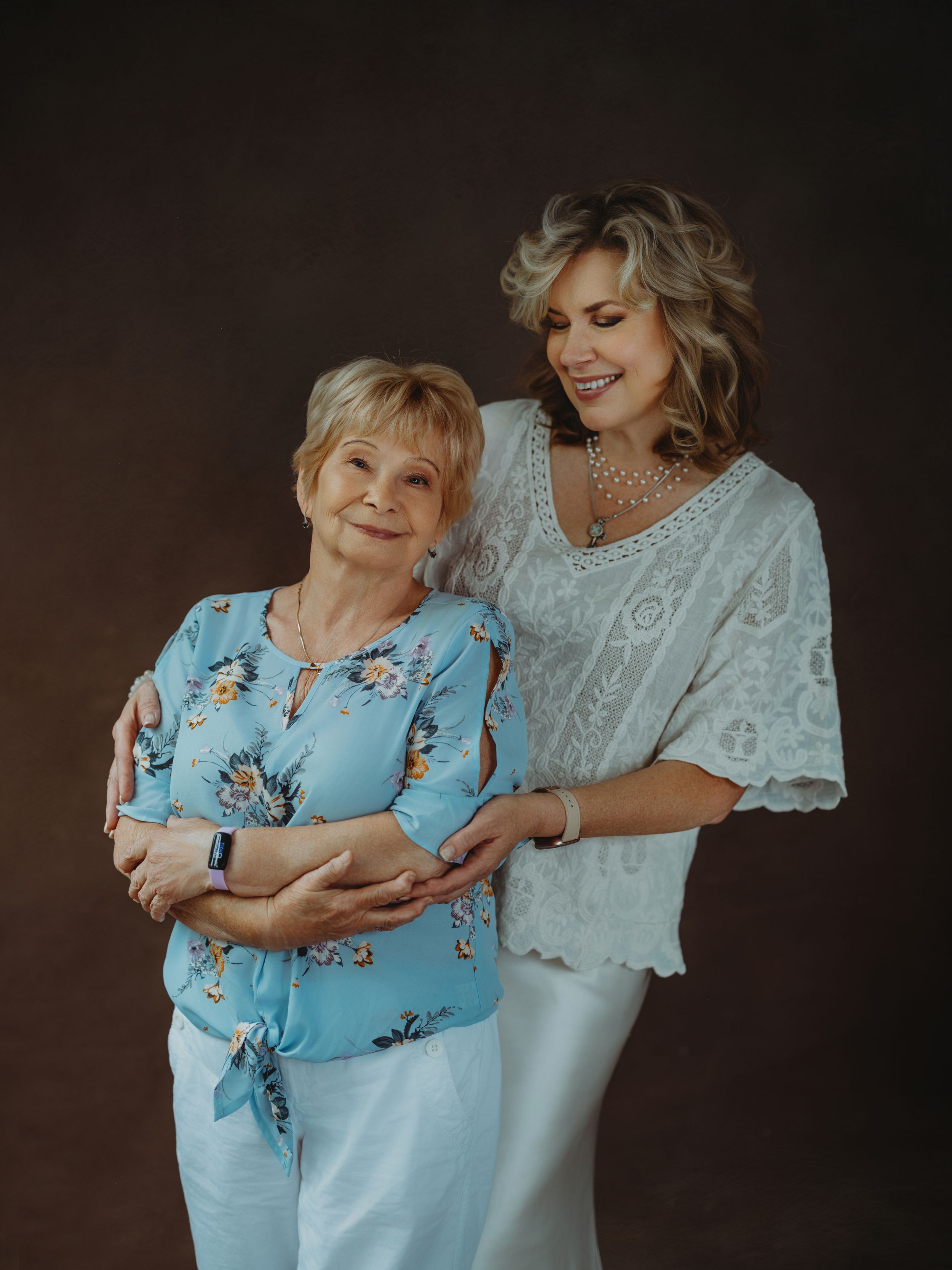 Two women in light colored outfits pose together against a dark backdrop in a studio portrait setting.