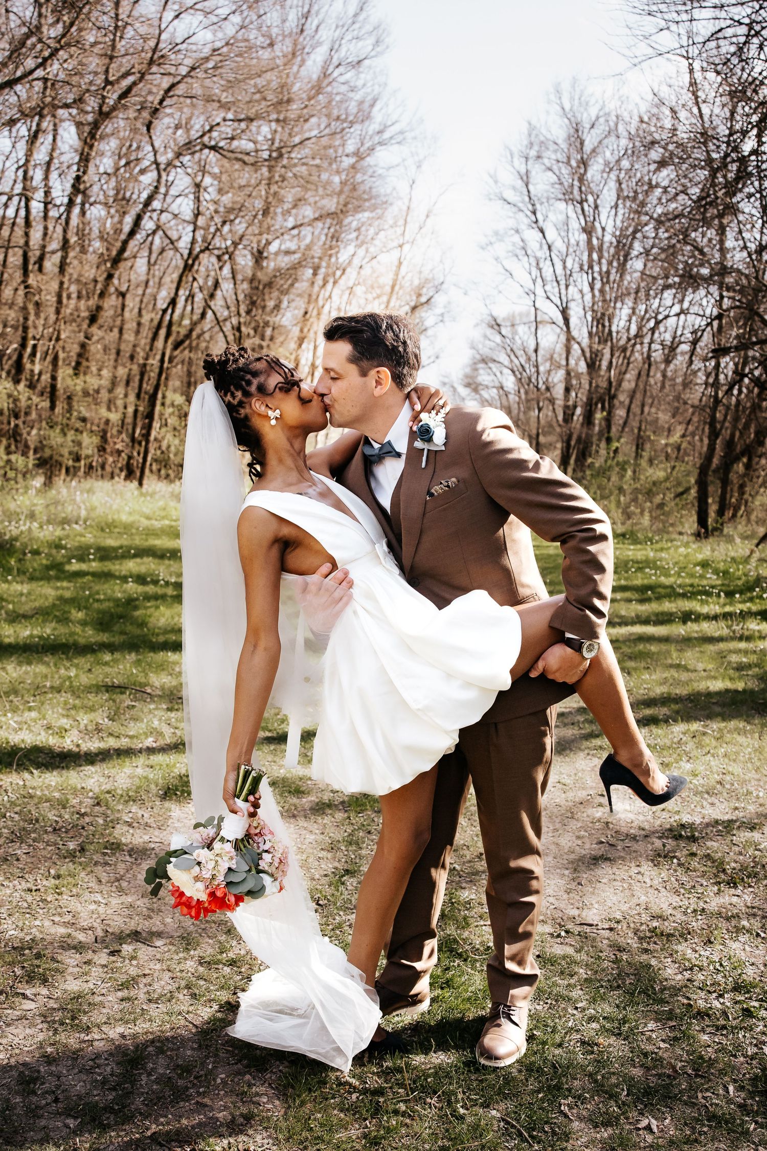 Groom in brown suit lifts and kisses bride in white dress outdoors among bare trees in spring setting.