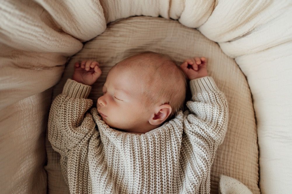 A newborn baby wearing a cozy knit sweater lies peacefully in a cream-colored bassinet.