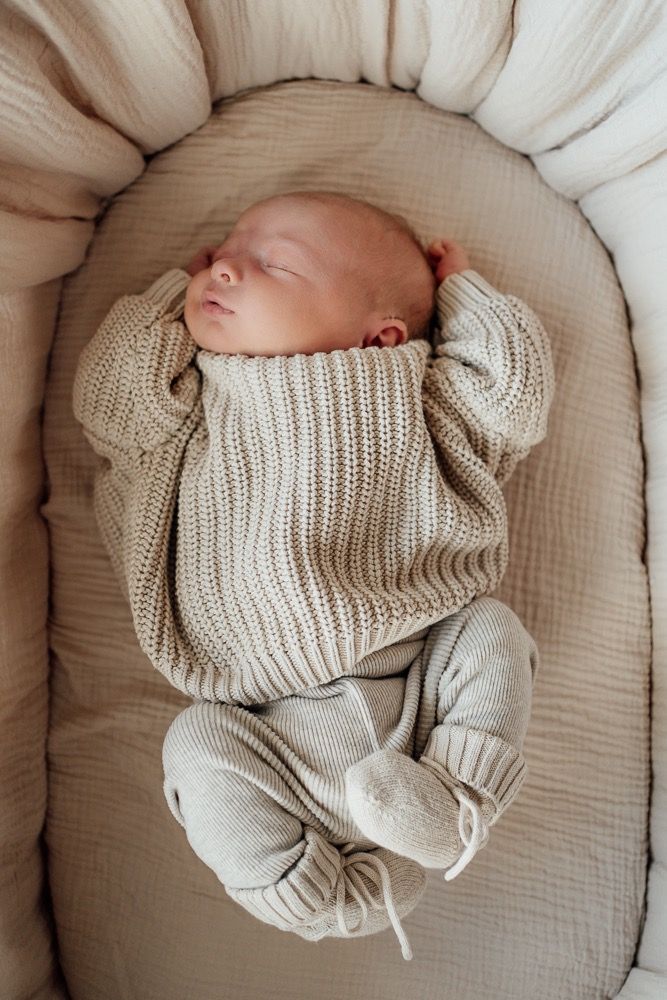 Sleeping newborn baby wearing cozy knit sweater lying on beige textured bedding.