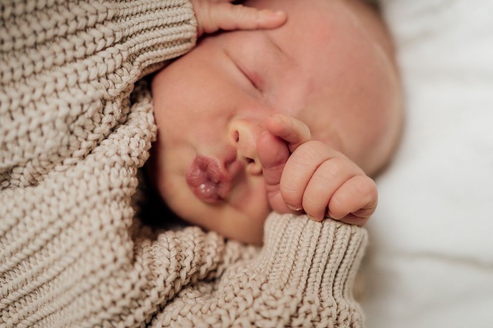 Close-up of a baby's tiny hand grasping a knitted beige sweater while sleeping on white bedding.